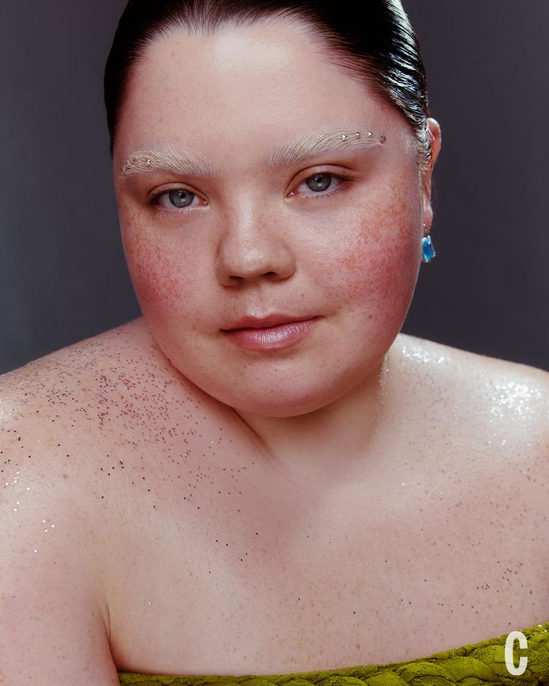 Close-up portrait of a woman with freckles, bleached eyebrows, and glitter dusting her shoulders against a plain grey backdrop.