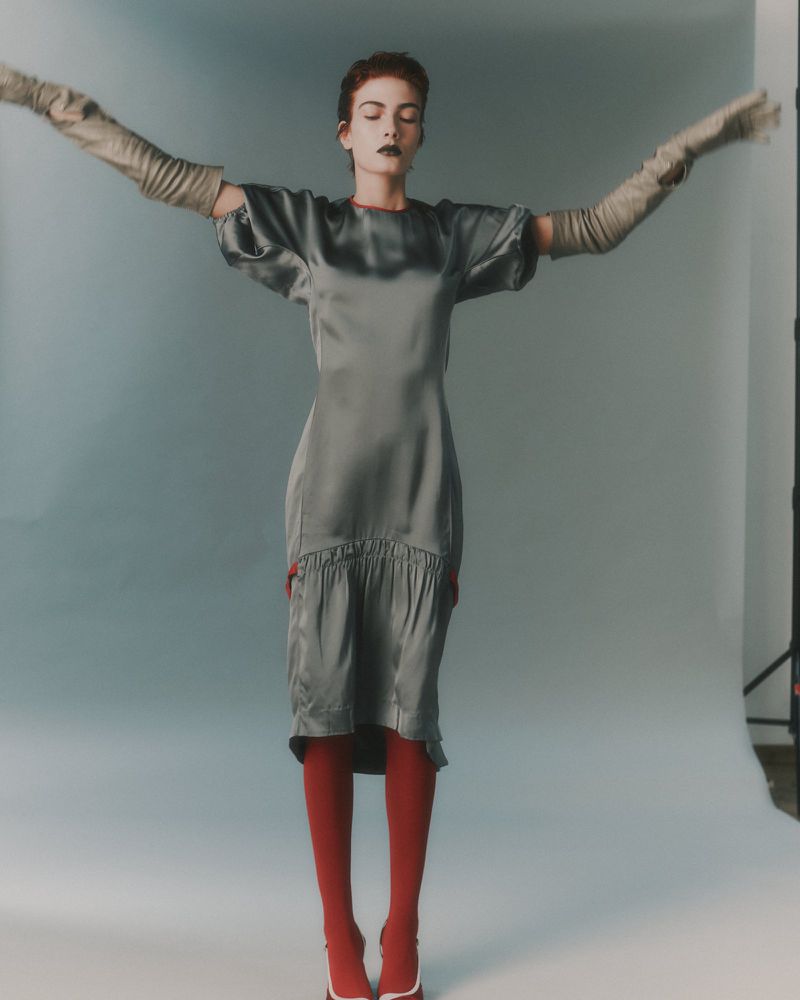 Model with arms outstretched wearing a satin midi dress, long grey gloves and red tights against a grey seamless backdrop