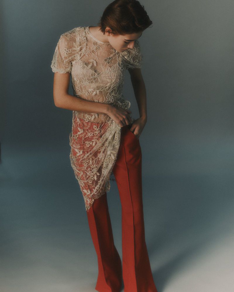 Model wearing a sheer lace top and red flare trousers against a gradient studio background.