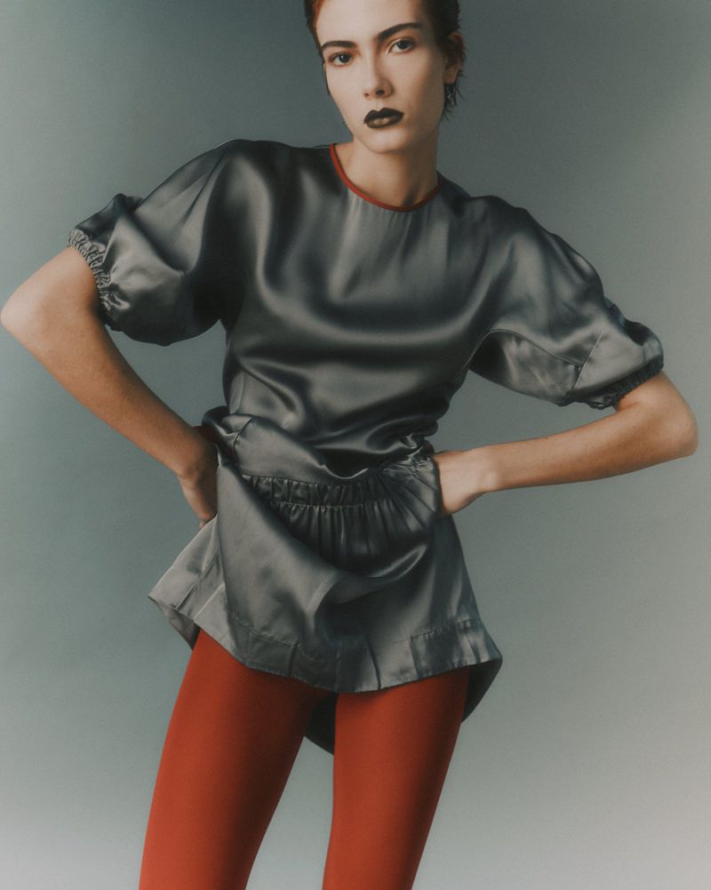 Model with dark lipstick wearing a grey satin top and red leggings against a gradient studio background