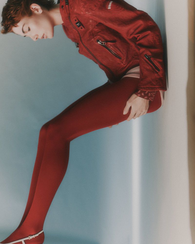 Model wearing a red leather jacket and red leggings against a blue and white studio background.
