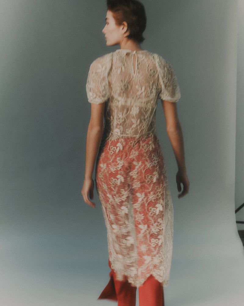 Back view of a woman with short brown hair wearing a sheer, floral lace top with puff sleeves and red pants in a neutral studio.