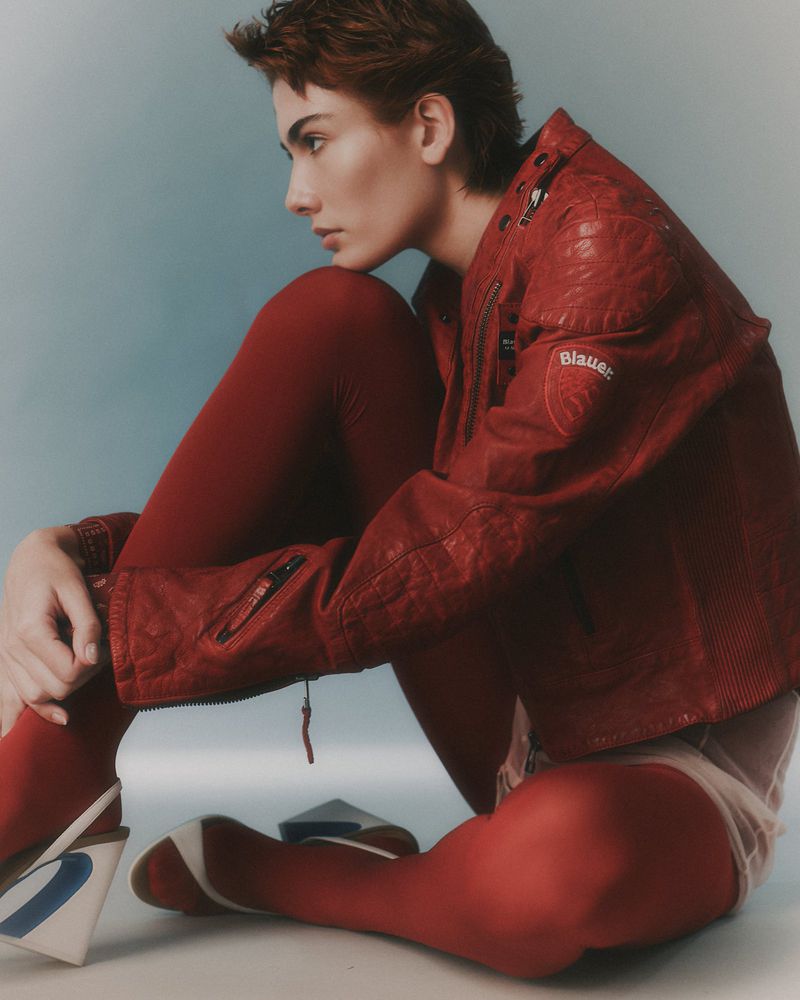 Model with short brown hair wearing a red leather jacket and red tights seated against a neutral studio background