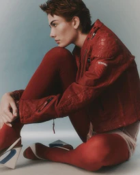 Model with short brown hair wearing a red leather jacket and red tights seated against a neutral studio background