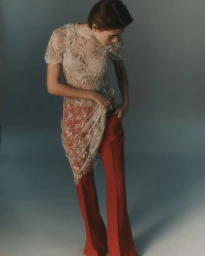 Model wearing a sheer lace top and red flare trousers against a gradient studio background.