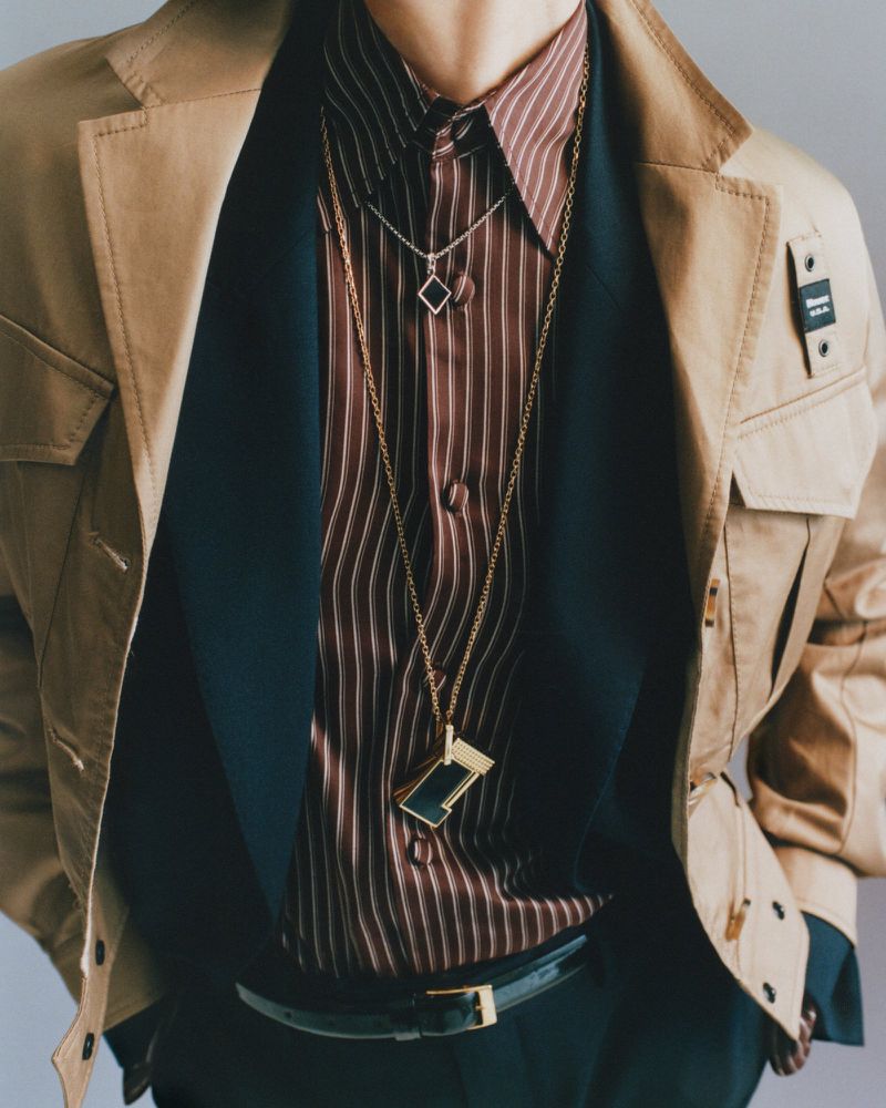 Close-up fashion editorial portrait of a model wearing layered jackets, a striped shirt, and gold pendant necklaces.