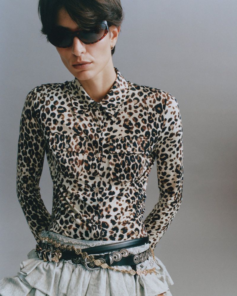 Model wearing a leopard-print shirt, dark sunglasses, and layered belts against a neutral grey studio background