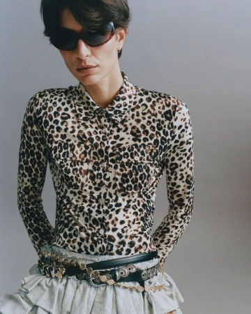 Model wearing a leopard-print shirt, dark sunglasses, and layered belts against a neutral grey studio background