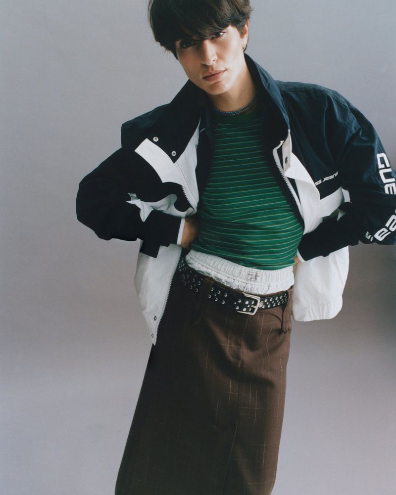 Model wearing a green striped top, black and white jacket, and brown skirt against a neutral grey background.