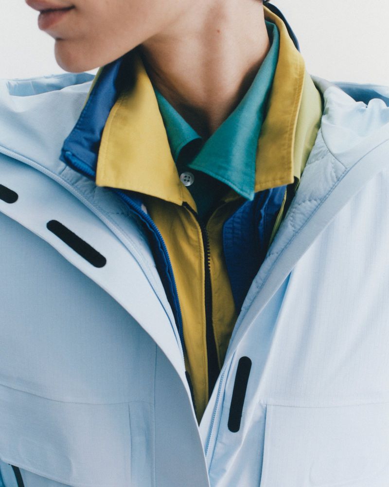 Close-up of a model wearing layered colorful shirts under a light blue jacket against a plain background
