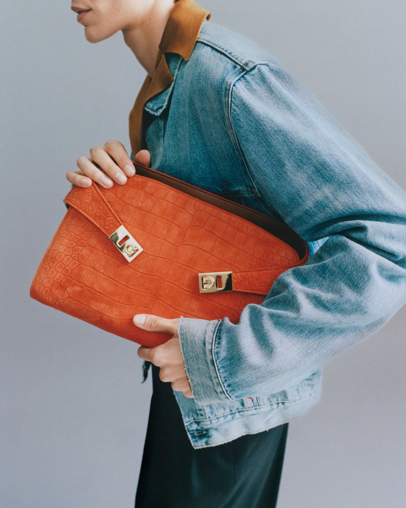 Model holding a vibrant orange textured leather clutch bag wearing a blue denim jacket against a neutral grey backdrop.