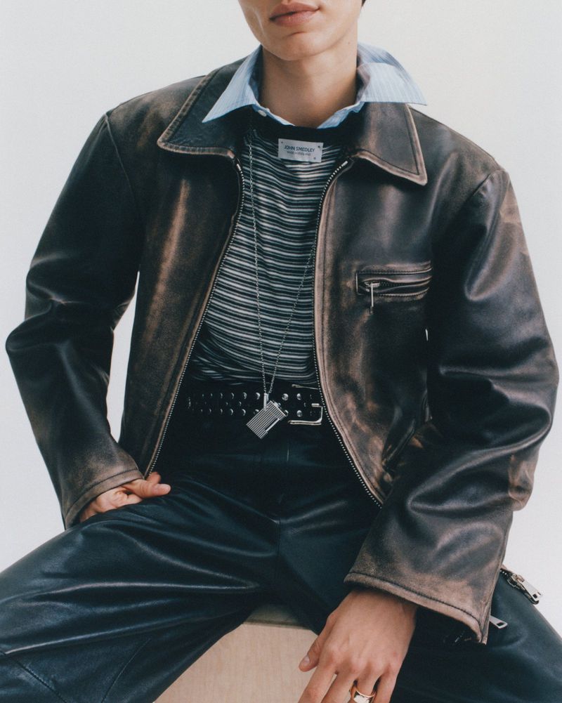 Model wearing a distressed leather jacket, striped top, blue shirt collar, and studded belt against a plain white background