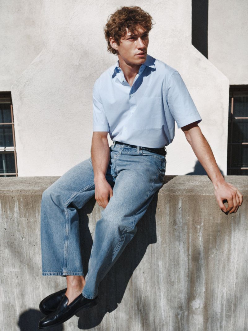 Male model seated on a concrete wall wearing a light blue short-sleeve shirt and blue jeans