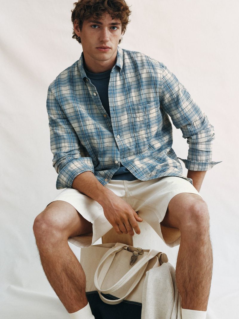 Model wearing a blue plaid shirt and white shorts seated above a canvas tote bag against a neutral background.
