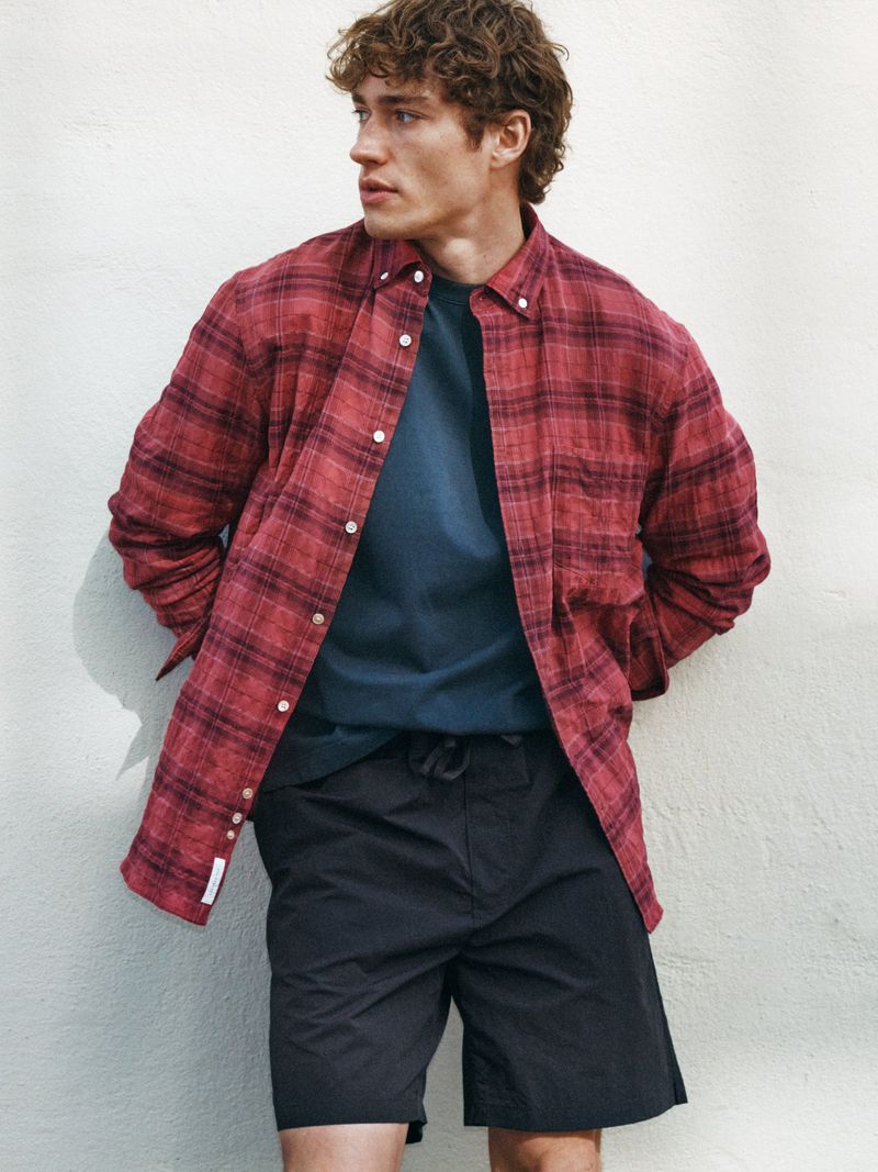 Model with curly hair wearing a red plaid shirt over a dark t-shirt and dark shorts against a textured off-white wall