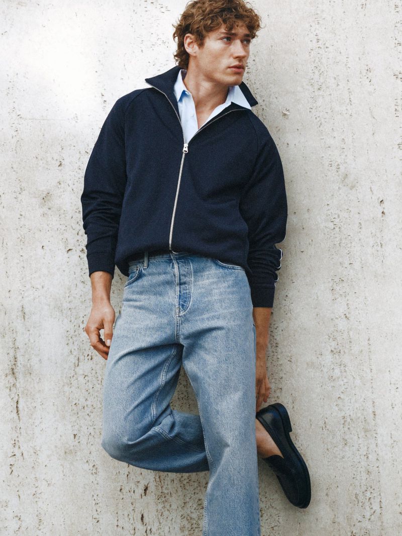 Model in dark blue zip jacket and light blue jeans leaning against a weathered concrete wall