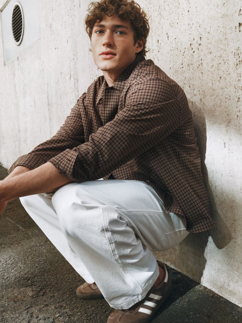 Model crouching against a textured stone wall wearing a brown checked shirt and white trousers