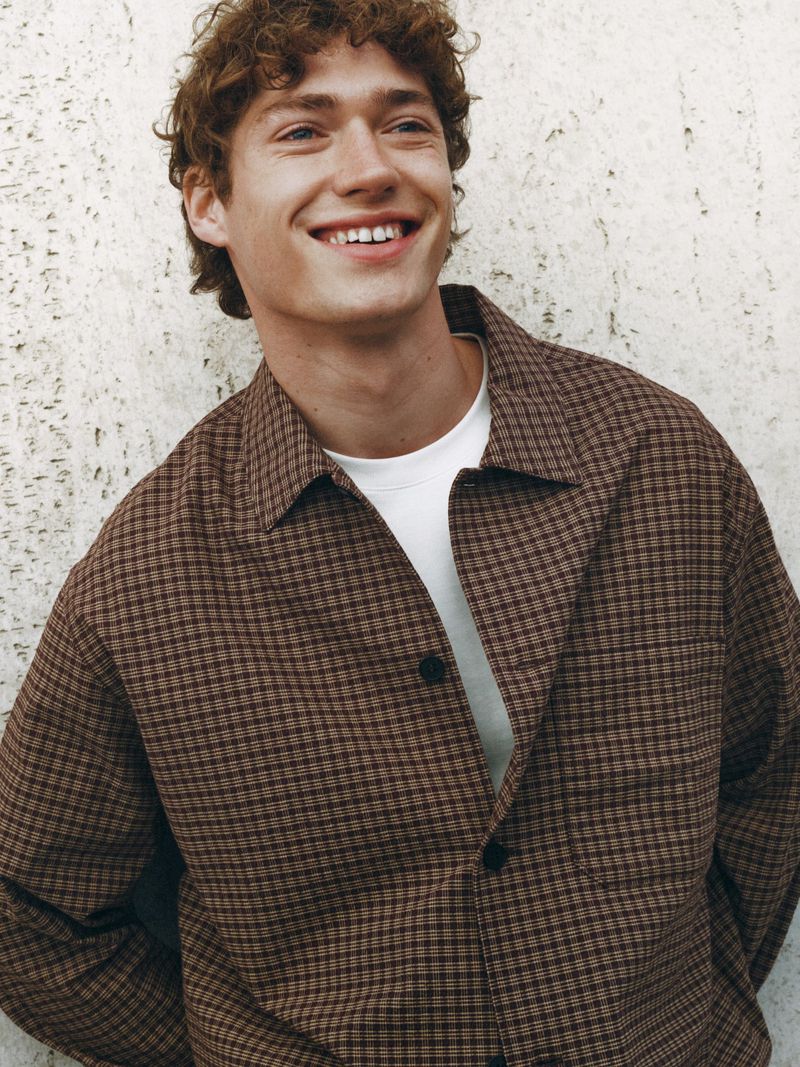 Smiling young man wearing a brown plaid overshirt and white t-shirt against a textured concrete wall.