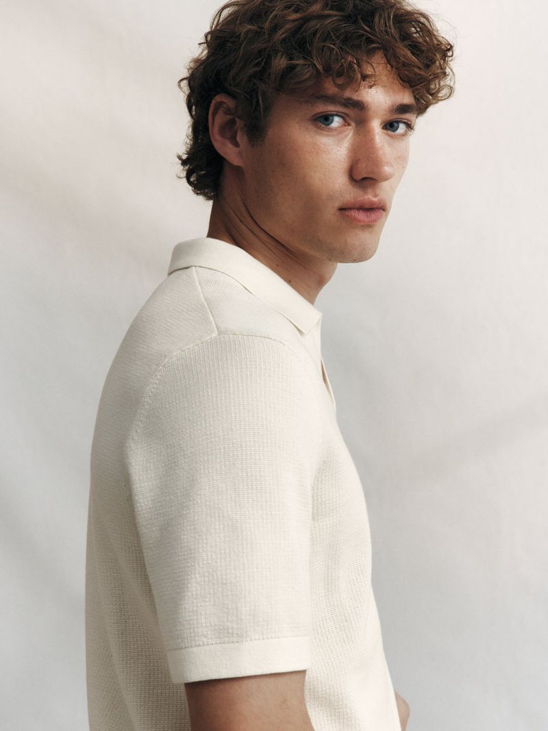 Young man with curly brown hair wearing a textured white polo shirt against a light studio backdrop