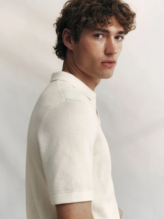 Young man with curly brown hair wearing a textured white polo shirt against a light studio backdrop