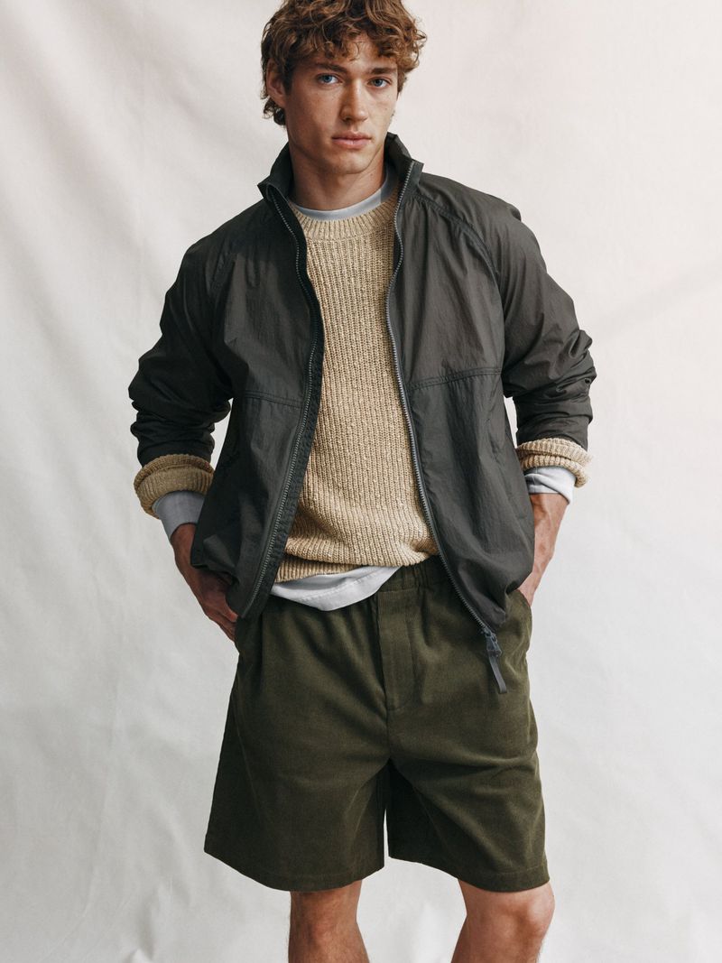 Male model in a dark zip jacket, textured beige sweater, and dark shorts against a seamless white studio backdrop