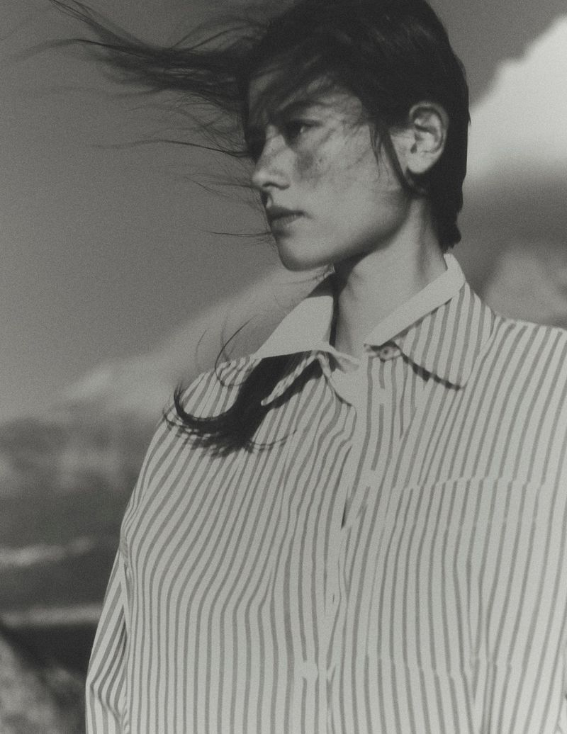 Model with dark hair blowing in the wind wearing a striped button-down shirt against an outdoor backdrop