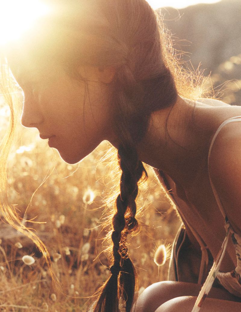 Close-up profile shot of a woman with a braid in a sunlit, grassy field at golden hour. Warm colors, peaceful scene, natural light.