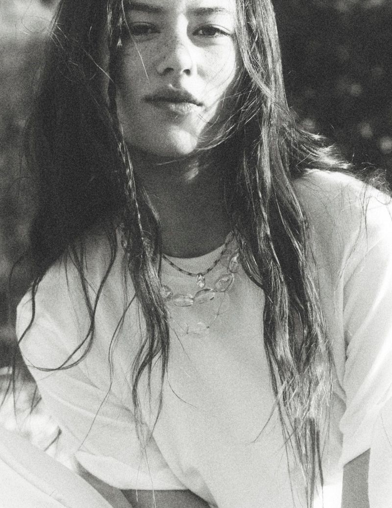 Portrait of Mahalia Celeste Henderson in a white t-shirt wearing layered beaded necklaces outdoors in black and white