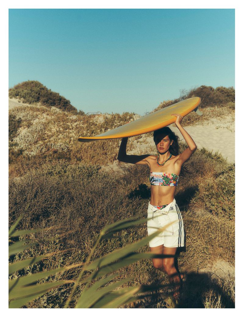 Woman carrying a yellow surfboard overhead standing among coastal sand dunes under a clear blue sky