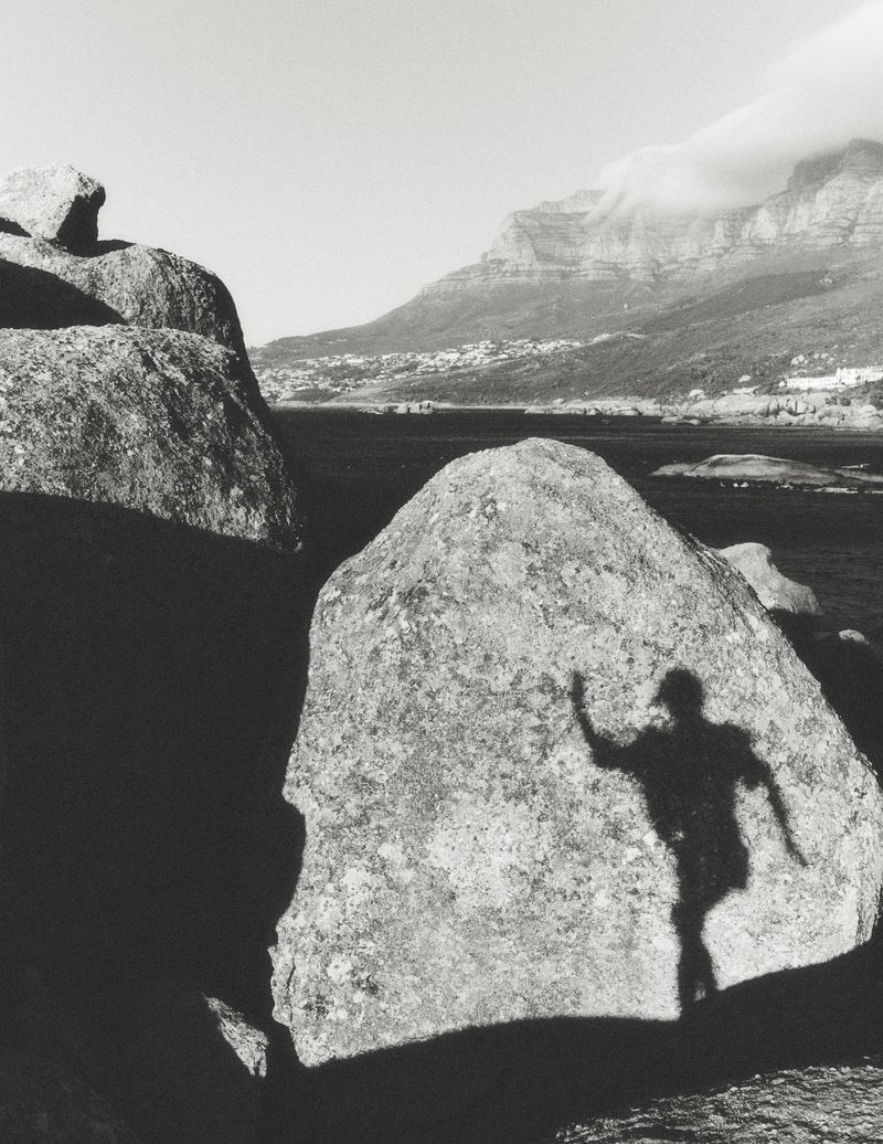 Black and white photograph of a human shadow cast onto a large boulder near the coast, with mountains in the background.