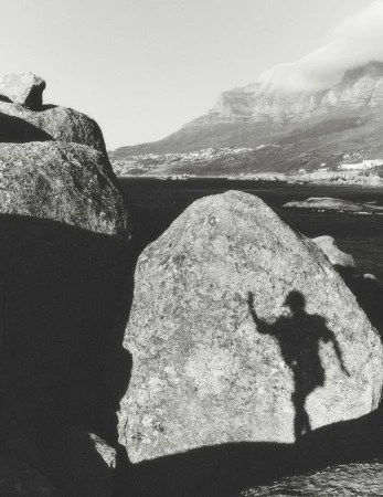 Black and white photograph of a human shadow cast onto a large boulder near the coast, with mountains in the background.