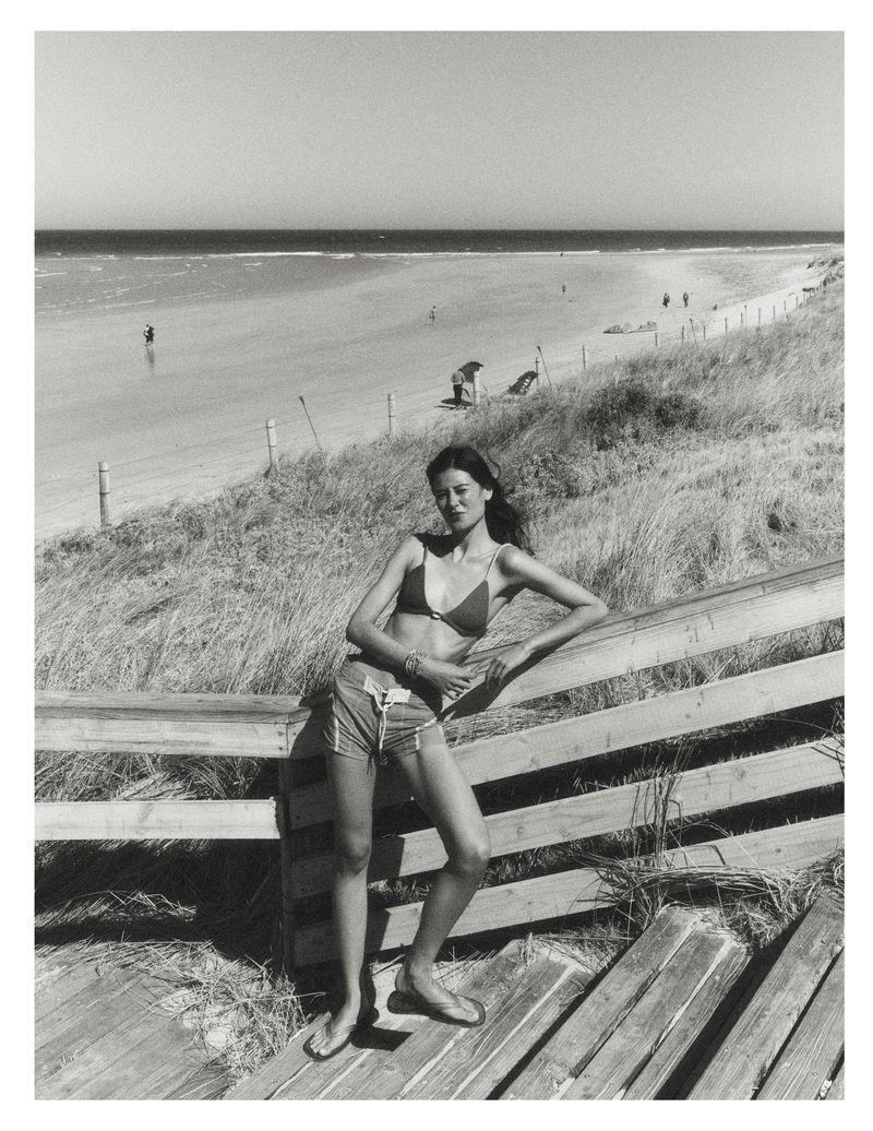 Model leaning against a wooden railing by a sandy beach and ocean, captured in black and white.