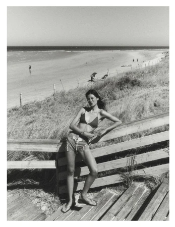 Model leaning against a wooden railing by a sandy beach and ocean, captured in black and white.