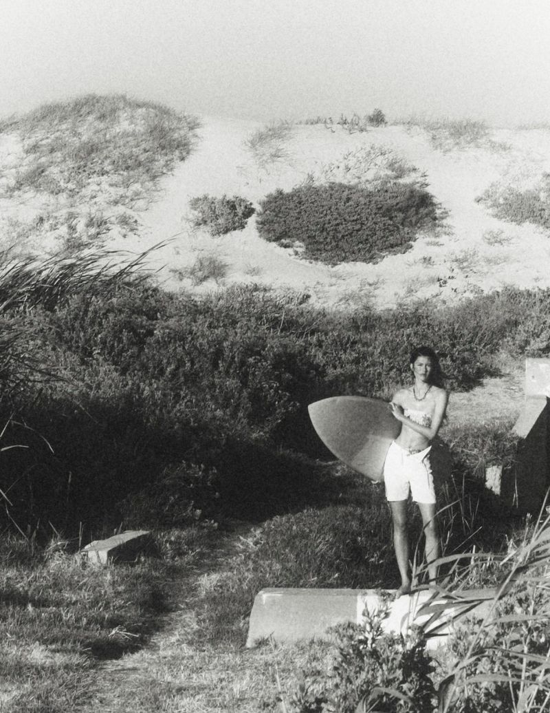 Model standing outdoors holding a surfboard near sand dunes and low vegetation in a black and white photograph