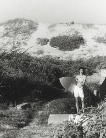 Model standing outdoors holding a surfboard near sand dunes and low vegetation in a black and white photograph