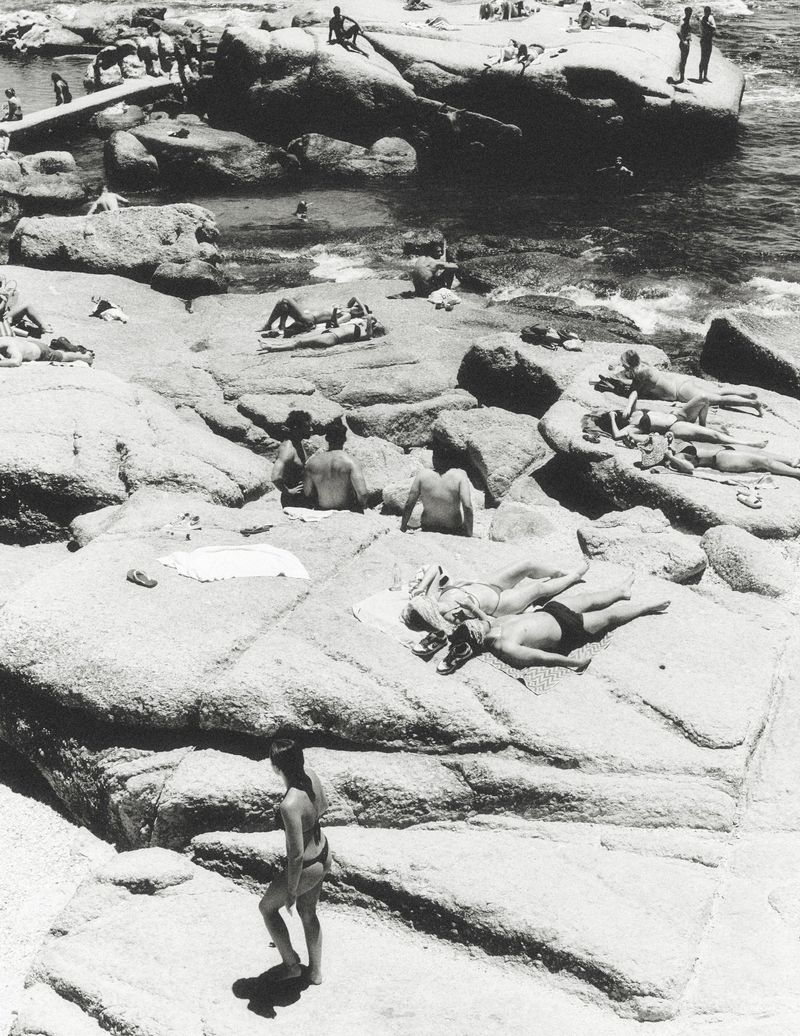 High-angle black and white photograph of people sunbathing and relaxing on large granite boulders near the ocean.