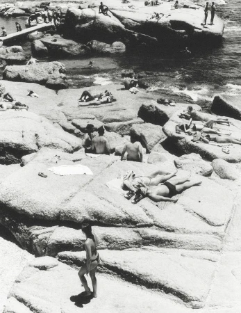 High-angle black and white photograph of people sunbathing and relaxing on large granite boulders near the ocean.
