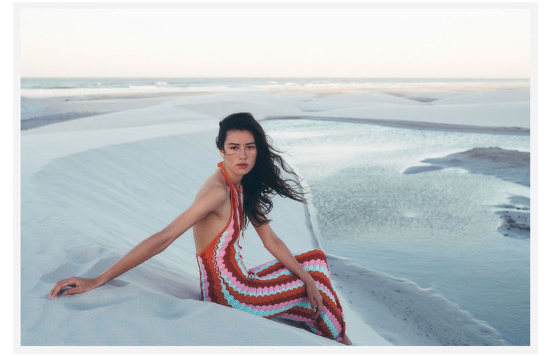 Model seated on white sand dunes by a calm coastal lagoon wearing a multi-colored zigzag halter dress for Anthropologie.