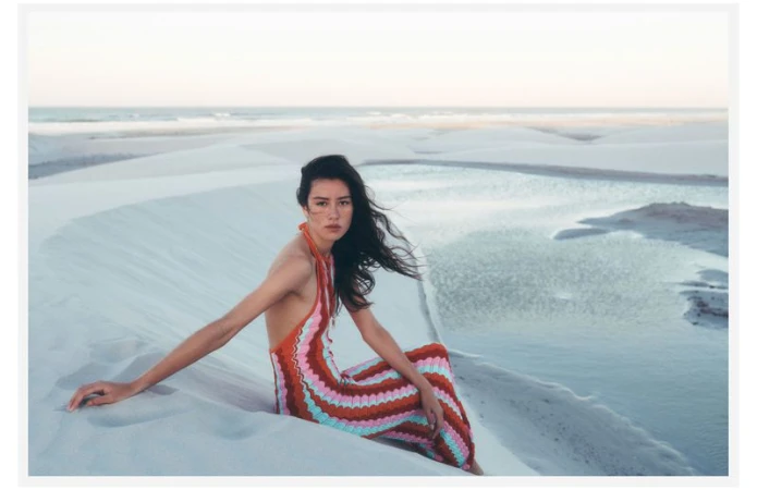 Model seated on white sand dunes by a calm coastal lagoon wearing a multi-colored zigzag halter dress for Anthropologie.