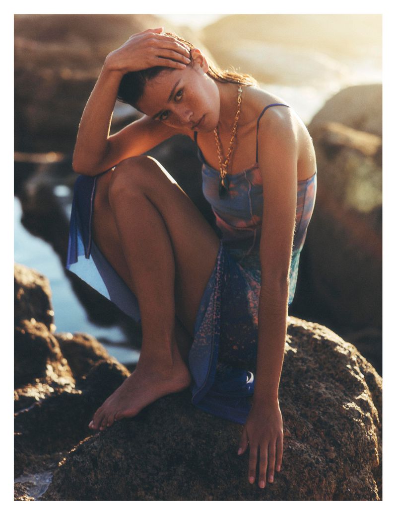 Model in blue slip dress crouching on rocky coastal shore during sunset for Anthropologie campaign