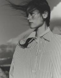 Model with dark hair blowing in the wind wearing a striped button-down shirt against an outdoor backdrop