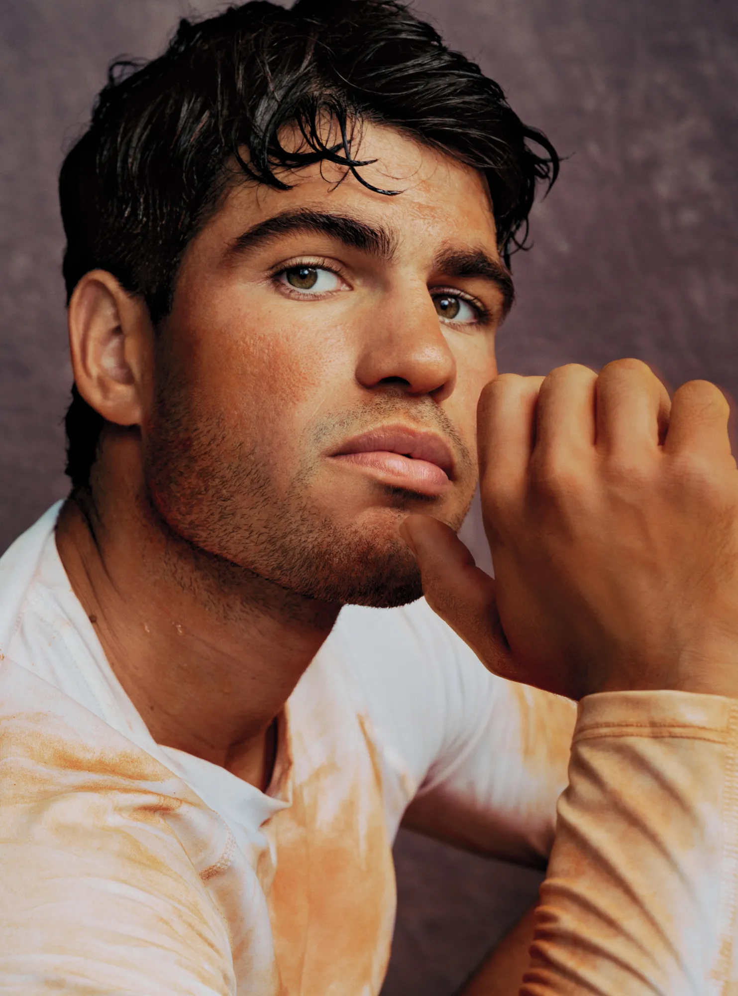 Close-up portrait of tennis player Carlos Alcaraz resting his chin on his hand, wearing a stained white top against a textured dark background.