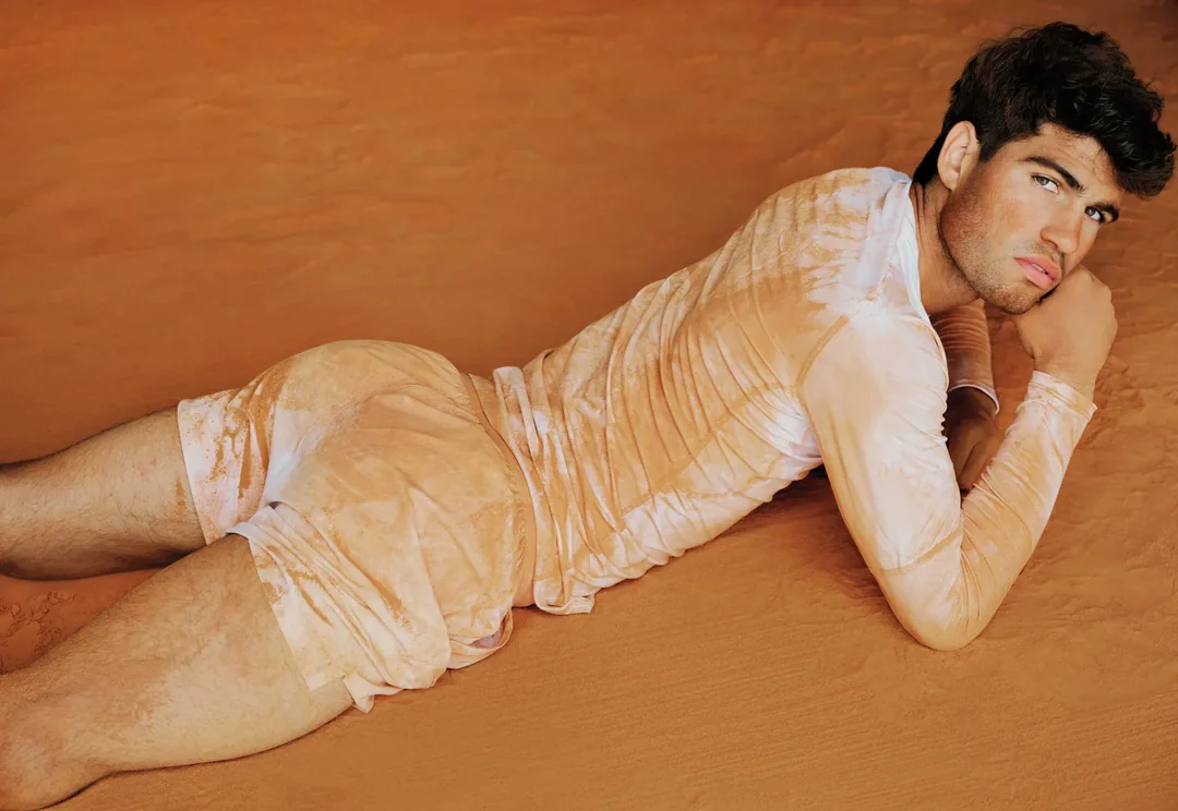 Carlos Alcaraz lying on a sandy surface wearing a cream patterned long-sleeve top and shorts for Vanity Fair U.S.