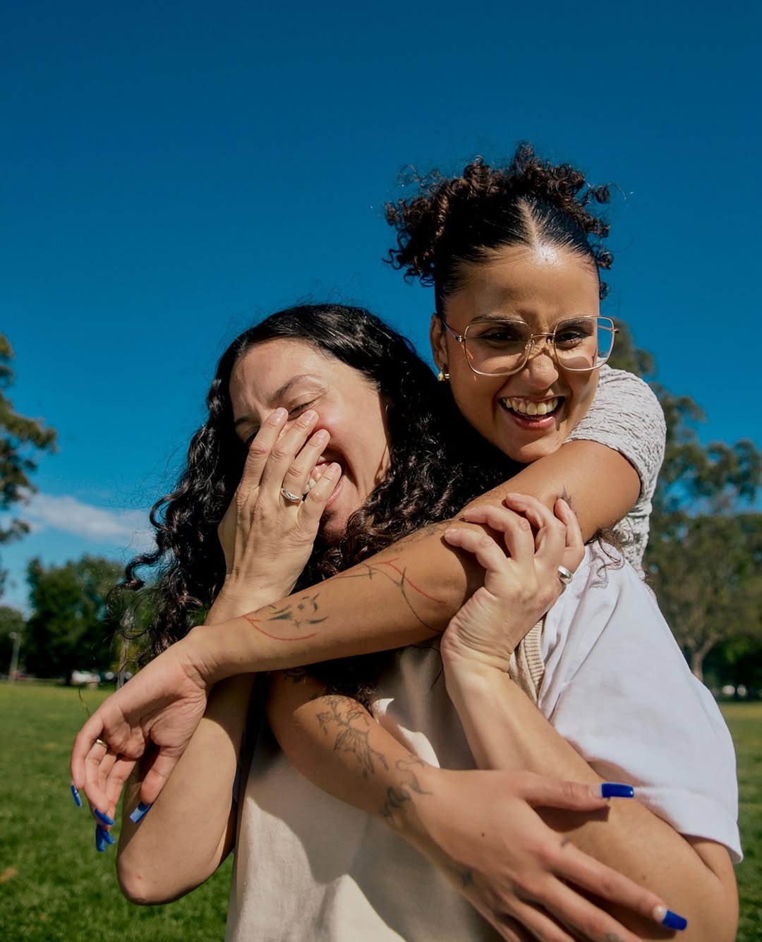 Two women embrace joyfully outdoors on a sunny day. One woman carries the other piggyback, both smiling with trees and blue sky behind.