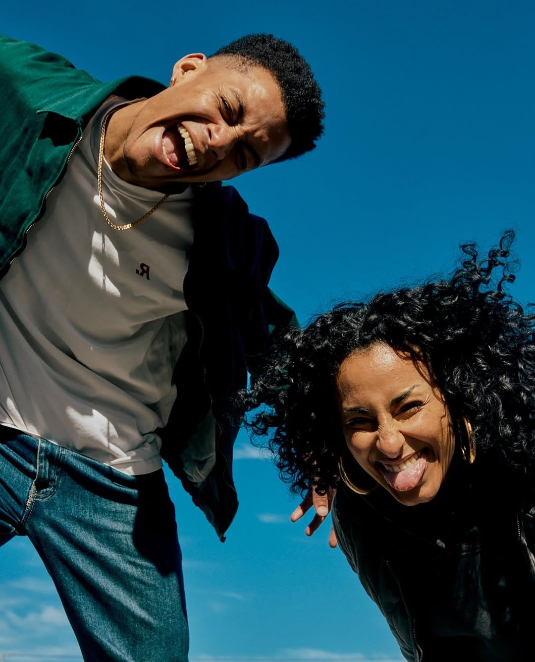 Two young adults pose outdoors against a bright blue sky, making silly faces and sticking out their tongues in a playful, low-angle shot.