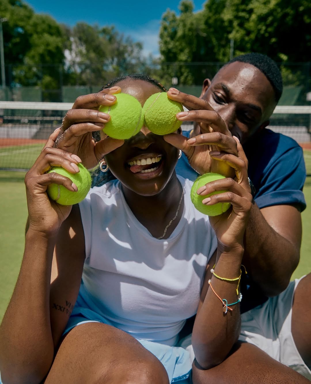 Playful tennis moment: A smiling African-descent woman holds tennis balls over her eyes, with an African-descent man behind her on a court.