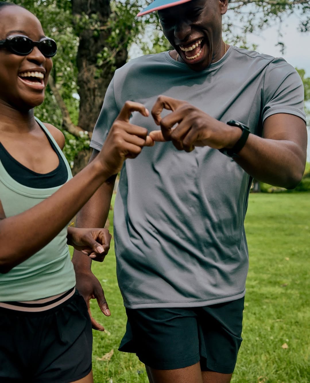 Two African American adults laugh and form heart shape with hands in an outdoor park setting, wearing casual athletic wear.