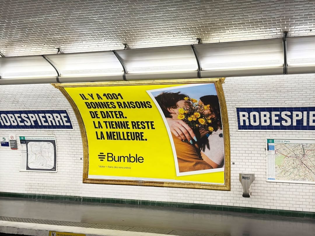 Bumble ad in a subway featuring a couple embracing with flowers, set against a bright yellow backdrop with French text and the Bumble logo.