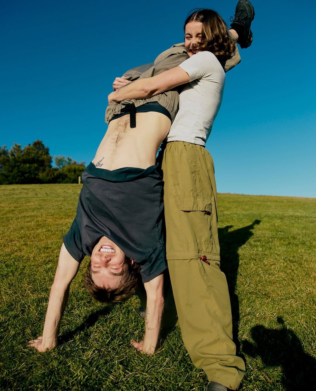 Woman holds man upside down on a grassy hill with blue sky. One adult female hugs a young adult male, bright daylight.