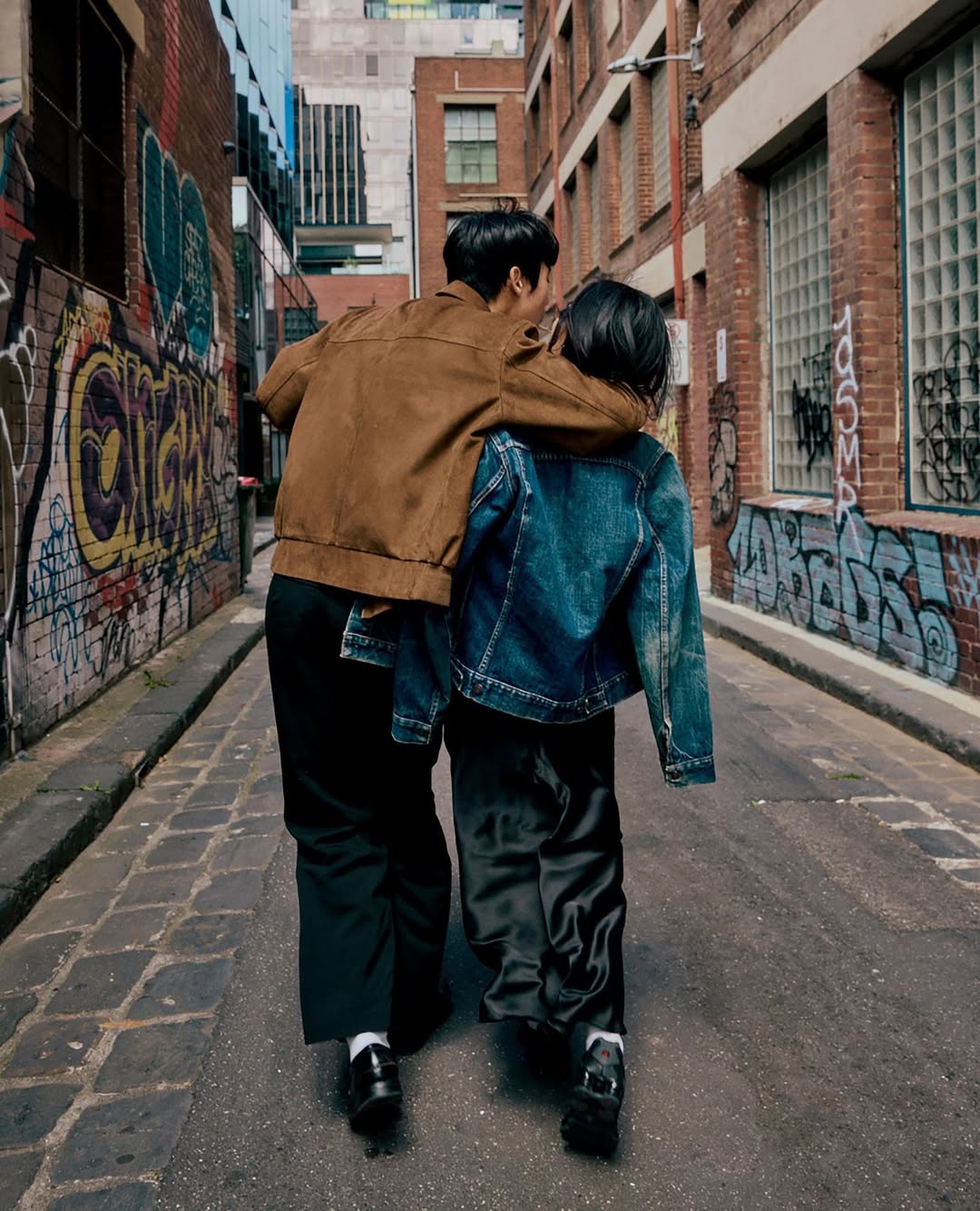 A couple walks down a city alleyway, arms around each other, wearing casual jackets and dark pants, framed by graffiti-covered brick walls.